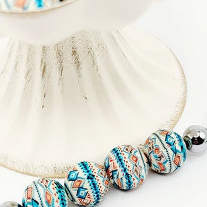 May include: A strand of colorful beads with a geometric pattern in shades of blue, orange, and white. The beads are strung on a silver cord.
