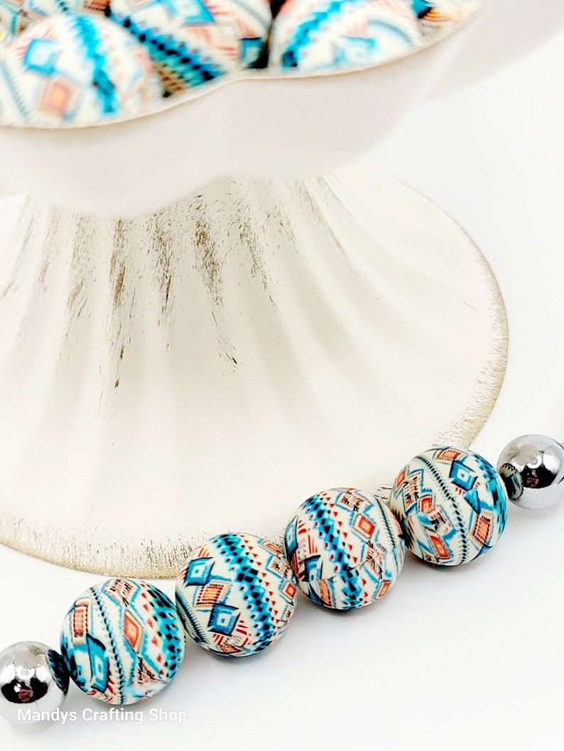 May include: A strand of colorful beads with a geometric pattern in shades of blue, orange, and white. The beads are strung on a silver cord.