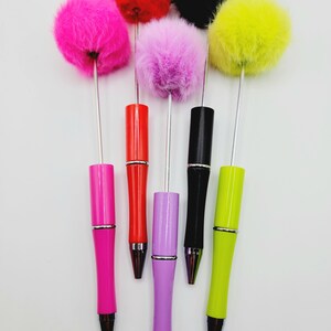 May include: Six colourful pens with fluffy pom-poms on top. The pens are pink, red, purple, black, yellow, and green.