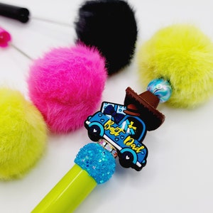 May include: A green pen with a blue truck charm that says "Best Dad" and a blue glitter accent. The pen is surrounded by four colourful pom-poms.