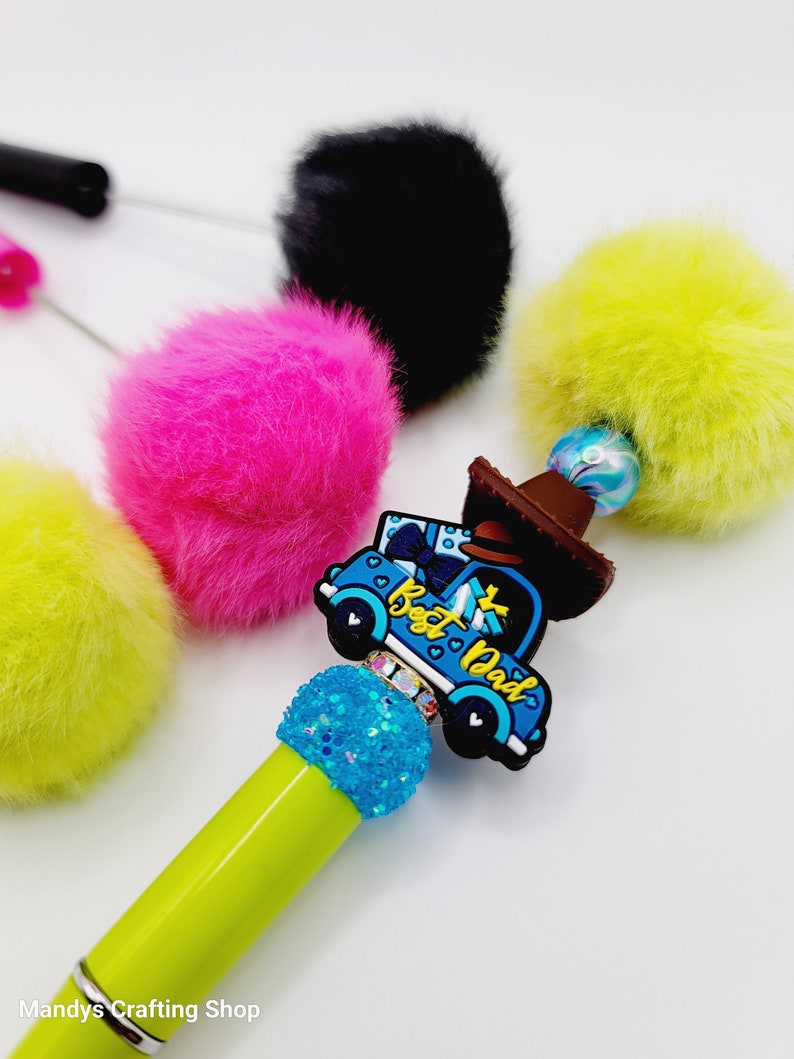 May include: A green pen with a blue truck charm that says "Best Dad" and a blue glitter accent. The pen is surrounded by four colourful pom-poms.
