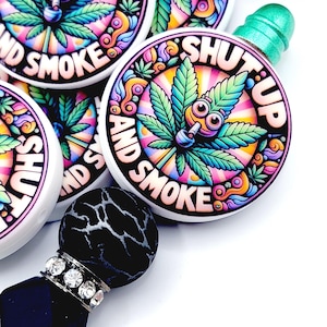 May include: A collection of colorful, round accessories with the text "SHUT UP AND SMOKE". Each accessory features a cartoon cannabis leaf with eyes and a smoking pipe, set against a vibrant, rainbow-colored background. The accessories have a white border.