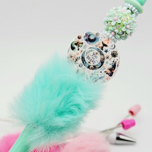 May include: A turquoise blue pen with a fluffy pom-pom top. The pen has a decorative top with a round charm that says "You are Brave" and a floral design with rhinestones.