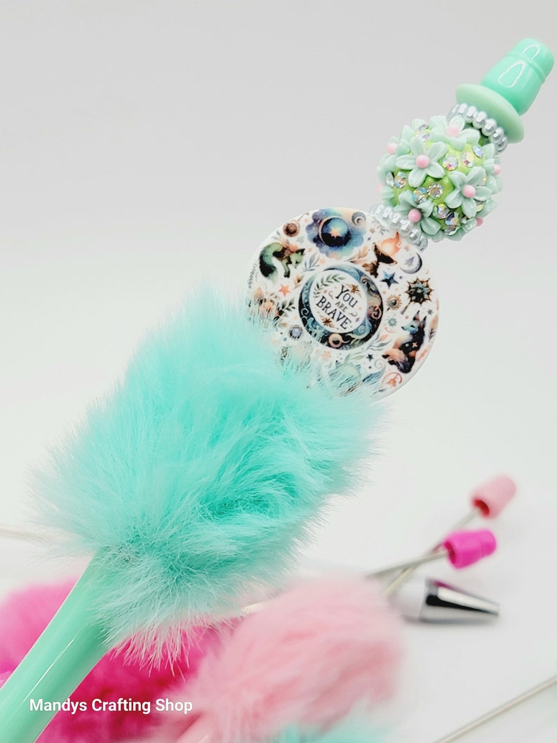 May include: A turquoise blue pen with a fluffy pom-pom top. The pen has a decorative top with a round charm that says "You are Brave" and a floral design with rhinestones.