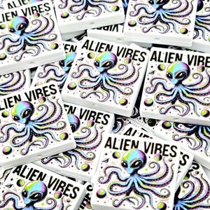 May include: White square-shaped beads with a colorful alien and octopus design. The text "ALIEN VIBES" is printed on each bead. The alien has a blue and green head with blue tentacles.