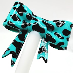 May include: A turquoise bow with black spots, mounted on a white stick. The bow has a glossy finish and a classic bow shape. The black spots are irregularly shaped, creating a playful pattern. The bow is a decorative accessory.