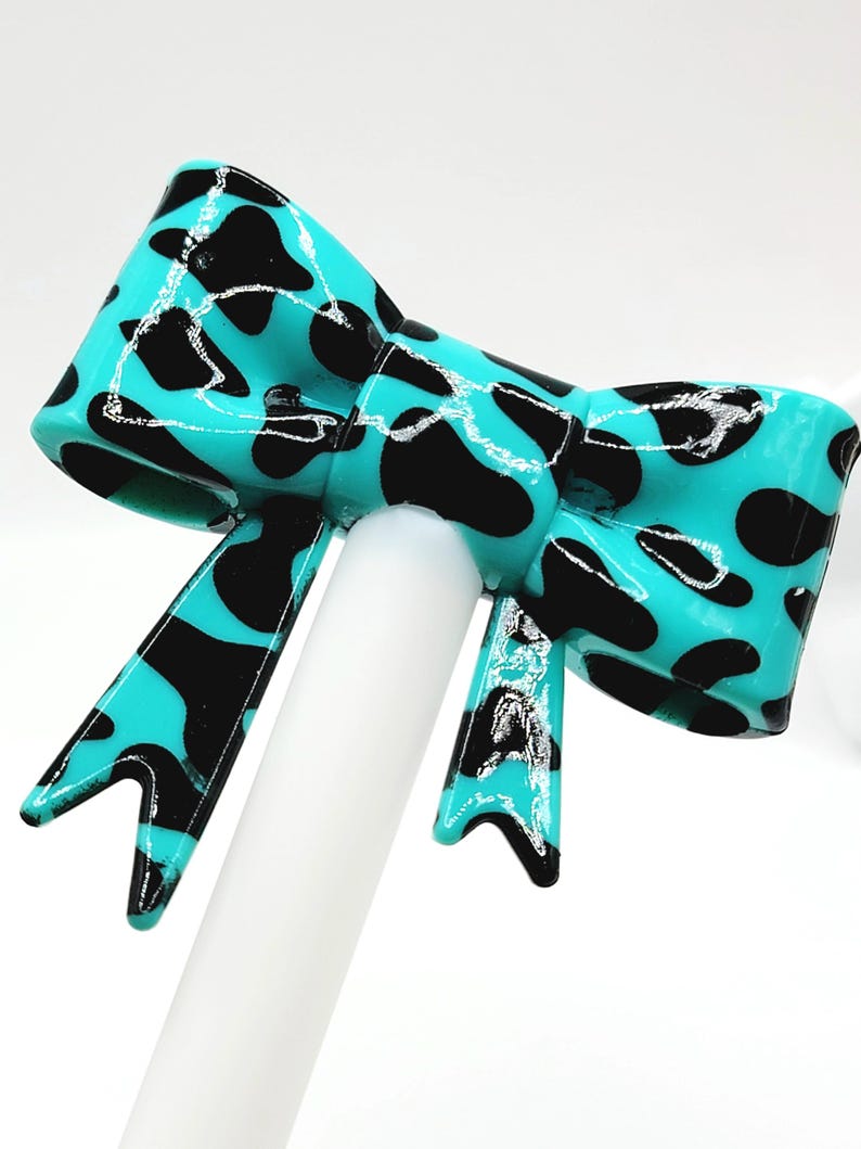 May include: A turquoise bow with black spots, mounted on a white stick. The bow has a glossy finish and a classic bow shape. The black spots are irregularly shaped, creating a playful pattern. The bow is a decorative accessory.