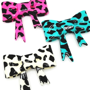 May include: Three glossy bow-shaped hair clips in a cow print design. One is bright pink with black spots, another is turquoise with black spots, and the third is white with black spots. The bows have a shiny finish and are set against a white background.