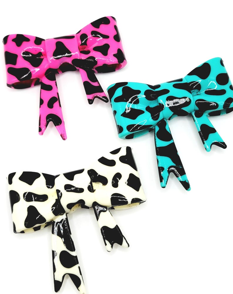 May include: Three glossy bow-shaped hair clips in a cow print design. One is bright pink with black spots, another is turquoise with black spots, and the third is white with black spots. The bows have a shiny finish and are set against a white background.