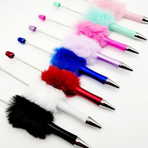 May include: A set of eight colourful pens with fluffy pom-poms on the top. The pens are arranged in a row from left to right, with the colours black, white, red, blue, purple, green, pink, and light pink.