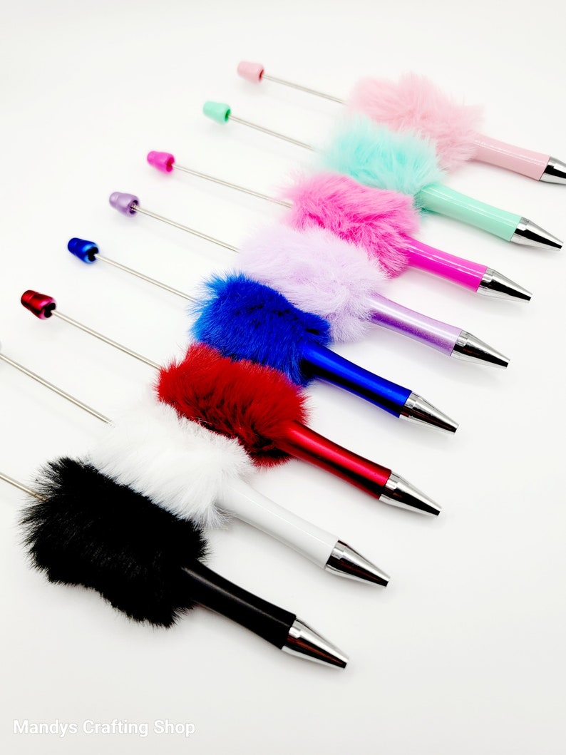May include: A set of eight colourful pens with fluffy pom-poms on the top. The pens are arranged in a row from left to right, with the colours black, white, red, blue, purple, green, pink, and light pink.