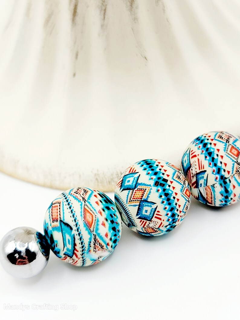 May include: Three round beads with a geometric pattern in shades of blue, orange, and white. The beads are strung on a silver metal connector.