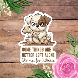 May include: A die-cut sticker featuring a grumpy, light brown and white puppy holding a large bone. The text reads: SOME THINGS ARE BETTER LEFT ALONE like me, for instance. The design has a white border and is set against a rustic wooden background.