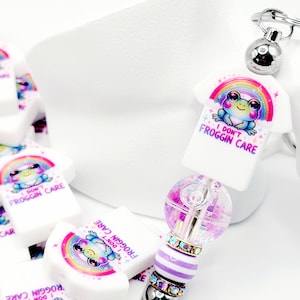 May include: A white charm with a rainbow and a frog wearing a shirt that says "I Don't Froggin Care". The charm is attached to a silver key chain with a purple and white striped bead and a clear bead with glitter.