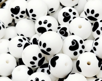 May include: A collection of white silicone beads with black paw print designs. The beads are round and have a hole in the center for stringing.