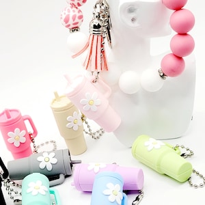 May include: A set of colorful silicone keychain charms shaped like a coffee mug with a daisy design. The charms are attached to silver chains. A pink silicone bracelet with a white and pink leopard print charm is also included.