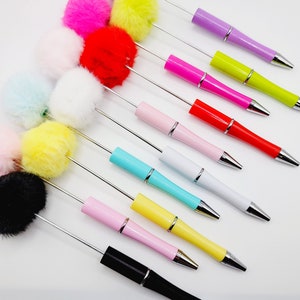 May include: A set of 12 colourful ballpoint pens with fluffy pom-poms on the top. The pens are arranged in a diagonal line, with the pom-poms facing upwards. The pom-poms are in various colours, including pink, yellow, white, blue, black, and red.