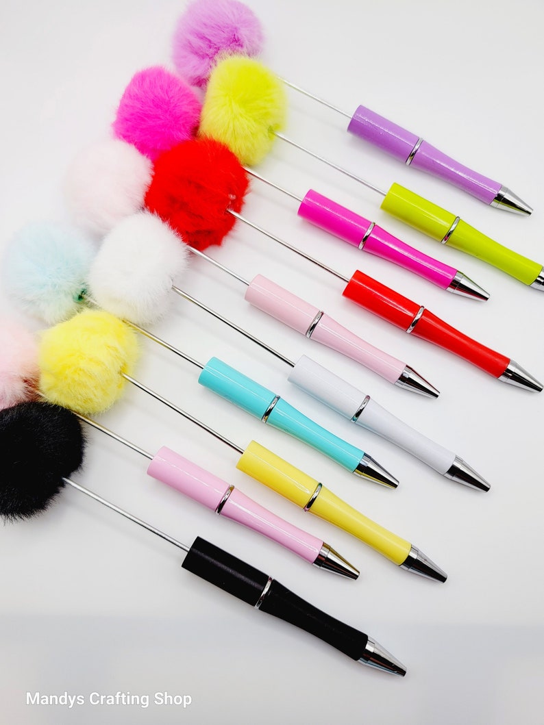 May include: A set of 12 colourful ballpoint pens with fluffy pom-poms on the top. The pens are arranged in a diagonal line, with the pom-poms facing upwards. The pom-poms are in various colours, including pink, yellow, white, blue, black, and red.