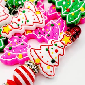 May include: A close-up of a Christmas themed charm bracelet with red and white striped beads, rhinestones, and colorful Christmas tree charms. The charm bracelet is made with a variety of colors, including red, white, green, pink, and yellow.