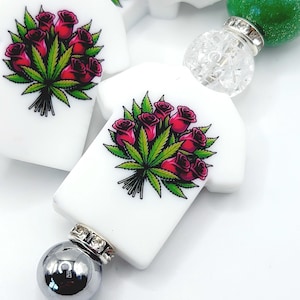 May include: White charm beads in the shape of a t-shirt, each featuring a graphic of red roses and green cannabis leaves. The beads are connected with silver and clear decorative elements, and a green bead.
