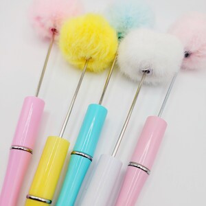 May include: Five colourful pens with fluffy pom-poms on the top. The pens are pink, yellow, blue, white, and light pink.