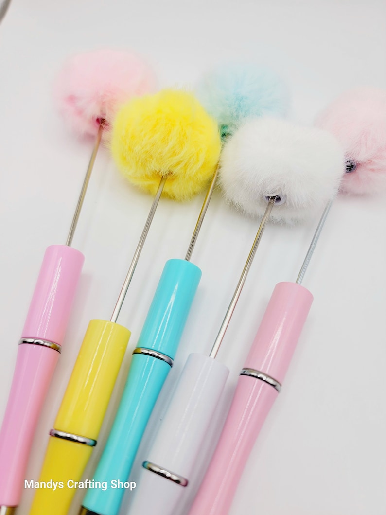 May include: Five colourful pens with fluffy pom-poms on the top. The pens are pink, yellow, blue, white, and light pink.