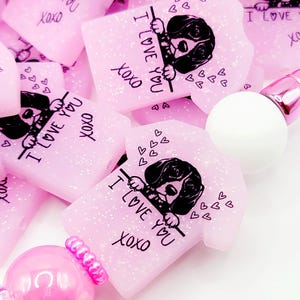 May include: Pink glitter resin charms shaped like t-shirts with a dog peeking over the top. The charms have the text "I love you xoxo" on them.