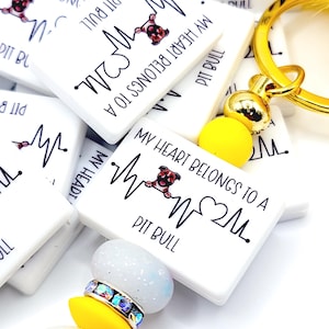 May include: White rectangular keychains with the text "MY HEART BELONGS TO A PIT BULL" and a heartbeat graphic. A gold keychain with yellow and white beads and a yellow fluffy pom-pom is in the foreground.