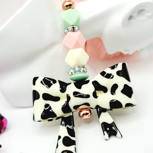 May include: A decorative keychain featuring a bow-shaped charm with a black and white cow print design. The keychain includes a gold-colored ring and a beaded strand with pastel-colored geometric beads and rhinestone accents. A small gold-colored ball is attached to the bow.