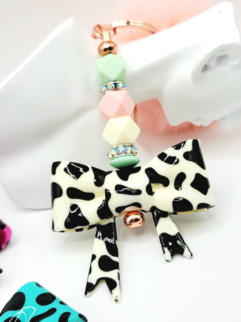 May include: A decorative keychain featuring a bow-shaped charm with a black and white cow print design. The keychain includes a gold-colored ring and a beaded strand with pastel-colored geometric beads and rhinestone accents. A small gold-colored ball is attached to the bow.