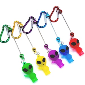 May include: A collection of colorful alien-shaped keychains. Each keychain features a different color alien figure, including green, yellow, purple, blue, and red, attached to a carabiner clip. The aliens have black eyes and are made of plastic.