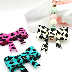 May include: Three bow-shaped accessories in pink, turquoise, and black and white cow print patterns. One is a keychain with a beaded accent in pastel colors. The bows have a glossy finish and are set against a white background.