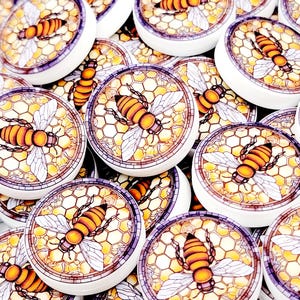 May include: A collection of round, white-edged bee-themed cabochons. Each cabochon features a detailed illustration of a bee with orange and black stripes, set against a honeycomb pattern. The cabochons are suitable for crafting projects.