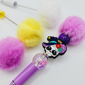 May include: Purple pen with a clear glitter ball and a sugar skull charm wearing a witch hat. The pen is surrounded by four fluffy pom-poms in white, yellow, and pink.