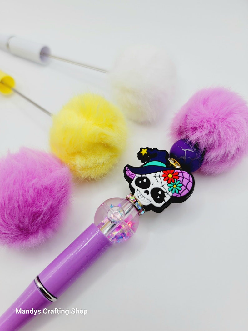 May include: Purple pen with a clear glitter ball and a sugar skull charm wearing a witch hat. The pen is surrounded by four fluffy pom-poms in white, yellow, and pink.