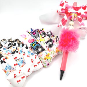 May include: A collection of colorful hair bows with various patterns, including cherries, leopard print, and floral designs. A pink pen with a fluffy pink pom-pom and a heart-patterned bow are also visible. The bows are arranged on a white surface.