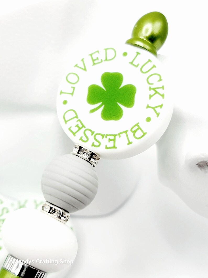 Peut inclure: Stylo blanc et vert avec un charme rond qui dit "Blessed. Loved. Lucky." avec un tr&egrave;fle &agrave; quatre feuilles au centre. Le charme est accentu&eacute; par des strass.