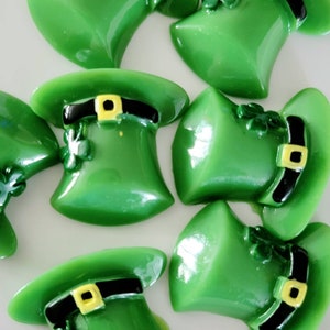 May include: Eight green leprechaun hats with black and yellow accents. The hats are made of a shiny, smooth material and have a three-dimensional design.