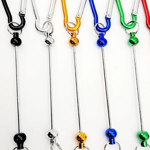 Op de afbeelding: Zes kleurrijke metalen sleutelhangers met een draaibare bal en een karabijnhaak. De sleutelhangers zijn zwart, zilver, goud, blauw, groen en rood.