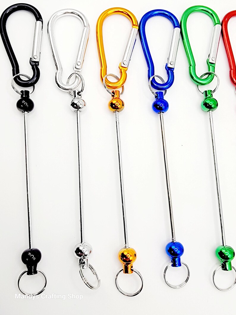 Op de afbeelding: Zes kleurrijke metalen sleutelhangers met een draaibare bal en een karabijnhaak. De sleutelhangers zijn zwart, zilver, goud, blauw, groen en rood.