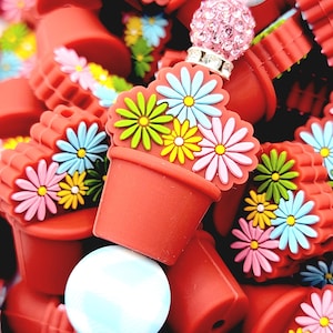 May include: A collection of red flower pot charms with colorful daisy accents. The charms are adorned with a variety of flower colors, including pink, yellow, blue, and green. The image also features a light blue and pink beaded charm.