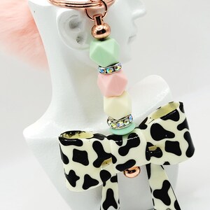 May include: A keychain with a rose gold ring and a pink fluffy pom-pom. The keychain has a beaded section with pastel green, pink, and yellow beads, and a cow print bow charm with a rose gold accent.