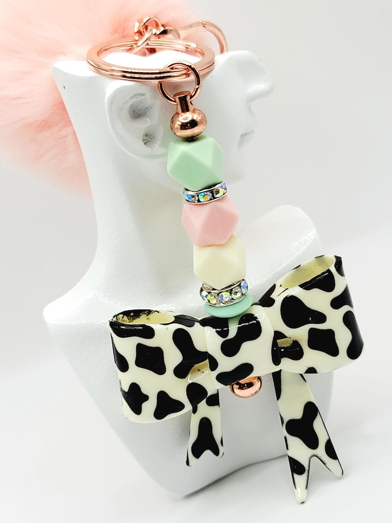 May include: A keychain with a rose gold ring and a pink fluffy pom-pom. The keychain has a beaded section with pastel green, pink, and yellow beads, and a cow print bow charm with a rose gold accent.