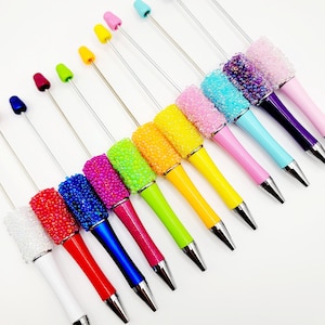 May include: A set of 12 colorful pens with jeweled tops. The pens are arranged in a diagonal line, with the jeweled tops facing up. The pens are all different colors, including white, red, blue, green, yellow, pink, and purple.