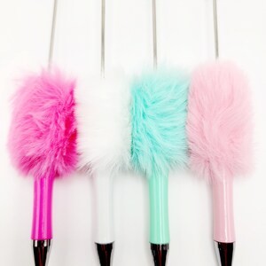 May include: Four fuzzy pens with different coloured fur tops. The pens are pink, white, light blue, and pink. The pens have silver tips and are on a white background.
