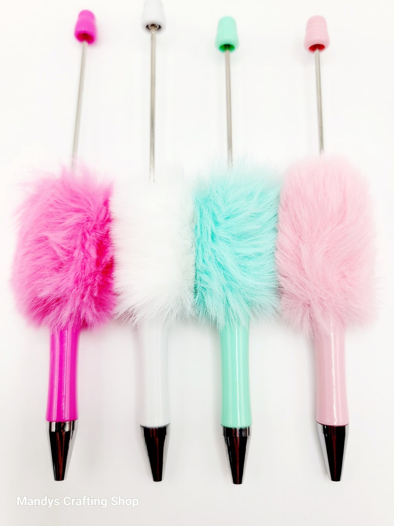 May include: Four fuzzy pens with different coloured fur tops. The pens are pink, white, light blue, and pink. The pens have silver tips and are on a white background.