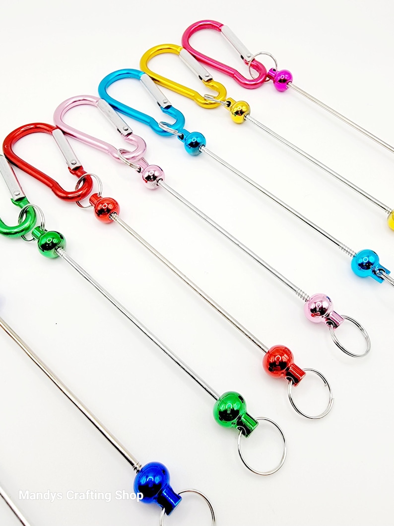 Op de afbeelding: Set van acht kleurrijke metalen sleutelhangers met een karabijnhaak en een metalen bal aan het einde van een lange metalen staaf. De sleutelhangers zijn in de kleuren rood, roze, blauw, groen, geel en paars.