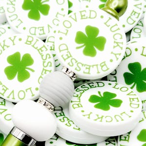 Peut inclure: Gros plan sur une collection de breloques rondes blanches avec des tr&egrave;fles &agrave; quatre feuilles verts et les mots "Blessed", "Loved" et "Lucky" imprim&eacute;s dessus. Les breloques sont dispos&eacute;es en pile, certaines se chevauchant. Au centre de l'image, il y a un stylo vert avec des perles blanches.