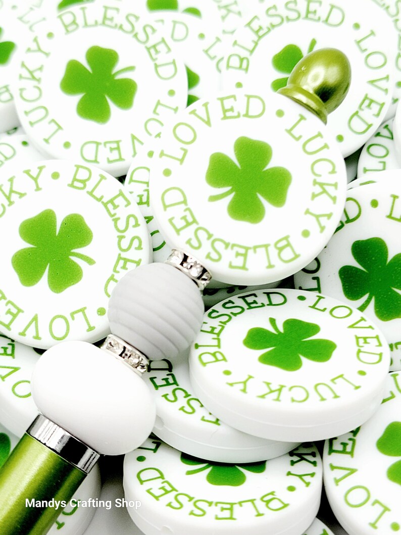 Peut inclure: Gros plan sur une collection de breloques rondes blanches avec des tr&egrave;fles &agrave; quatre feuilles verts et les mots "Blessed", "Loved" et "Lucky" imprim&eacute;s dessus. Les breloques sont dispos&eacute;es en pile, certaines se chevauchant. Au centre de l'image, il y a un stylo vert avec des perles blanches.