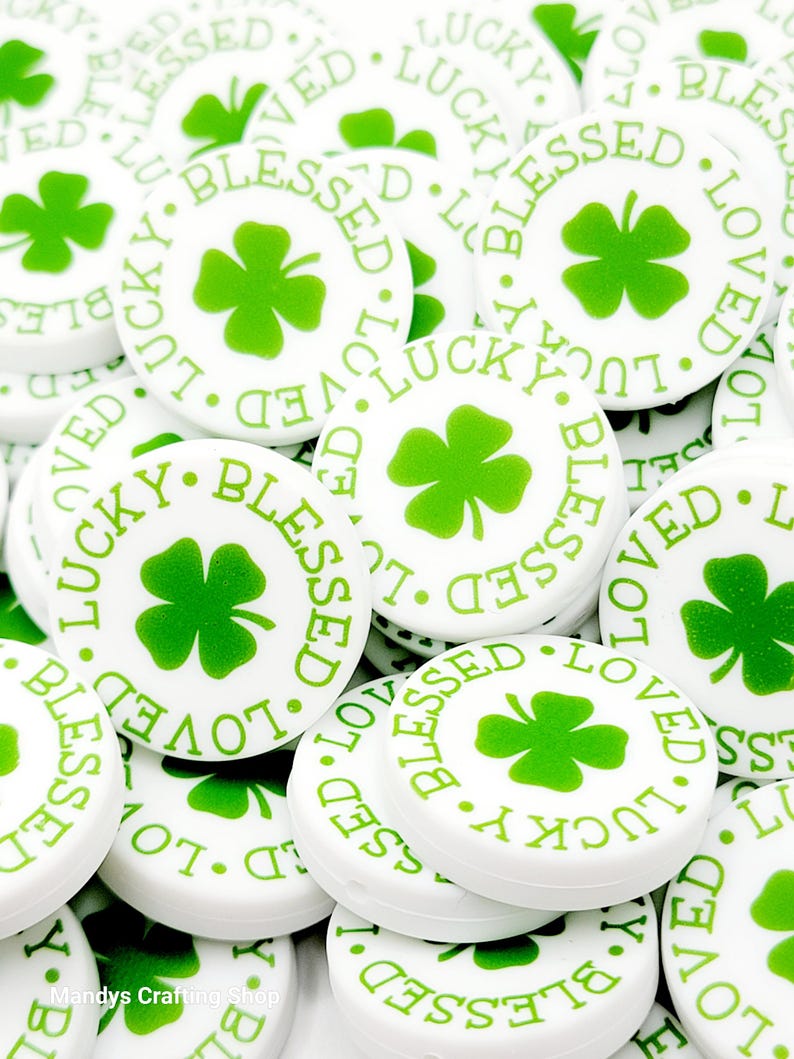 Peut inclure: Un tas de boutons ronds blancs avec des tr&egrave;fles &agrave; quatre feuilles verts et les mots "Lucky", "Blessed" et "Loved" imprim&eacute;s dessus.